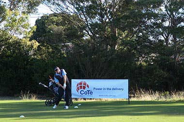 A golfer sets up to take a swing at the ball in front of a sponsor sign that reads Cote Power in the delivery