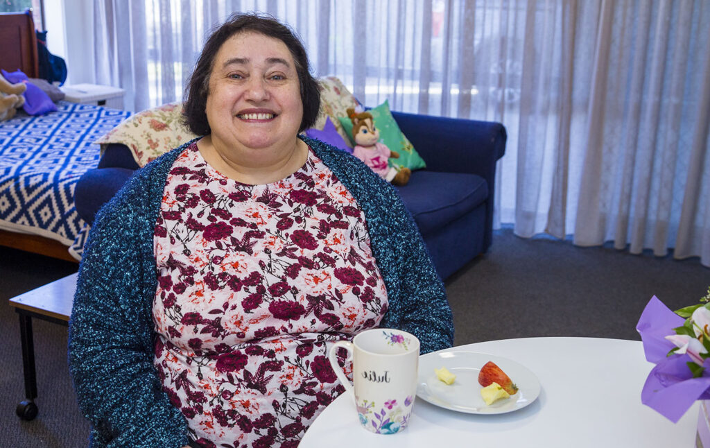 dark head lady sitting at table with a cup of tea and plate of food. She has a bed behind her.