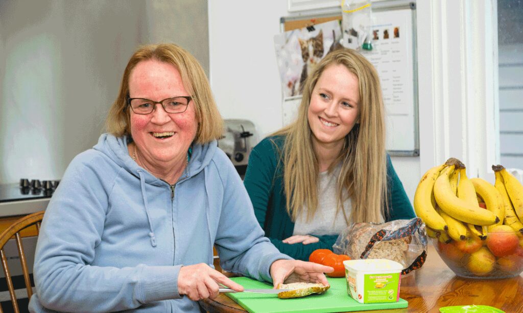 women smiling while buttering piece of bread with her specialist disability accomodation support worker next to her