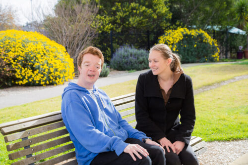 Young male participant Ben sitting on a park bench smiling with support worker next to his side