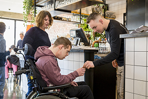 A person in a wheelchair hands money to a cafe worker. Another person stands beside the wheelchair and watches on