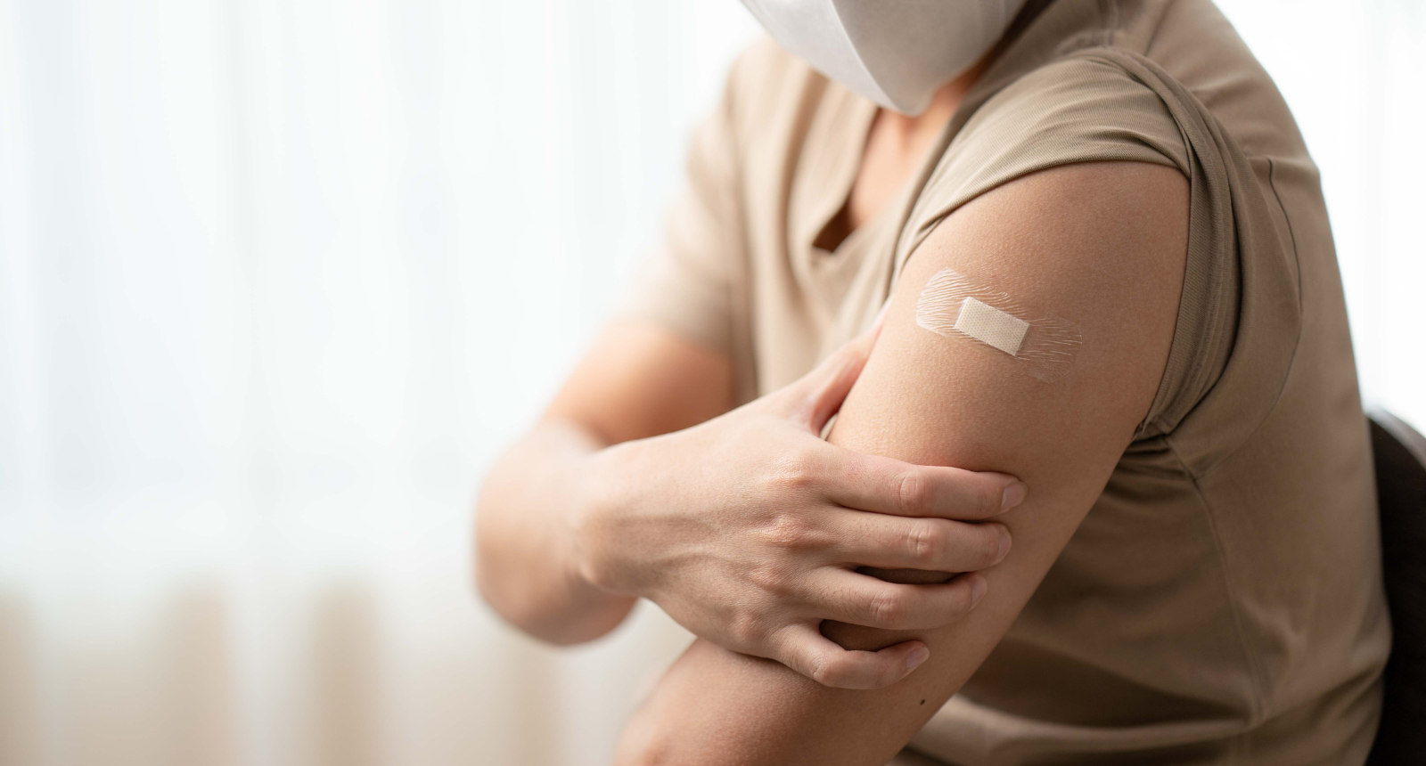 A person is sitting in a chair, holding their arm which has a bandaid on it to show they have been vaccinated.