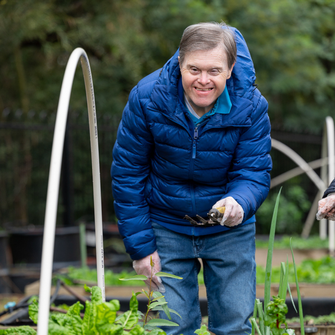 A person leans over a vegetable patch with a blue puffy jacket, holding a small spade and looking at the camera, smiling