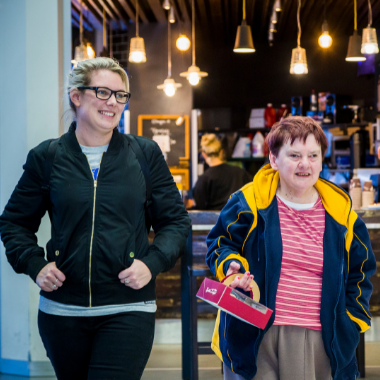 Two peple walking in a shopping centre. Both are smiling. One is wearing glasses and has her hands in her pockets. The second is carrying a parcel.
