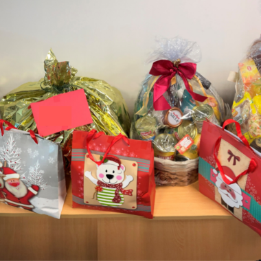 Three large Christmas hampers sit on a bench. In front are 3 Christmas gift bags