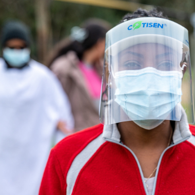 A person wearing a face guard and mask stares into the camera. To the left is a blurred image of a person wearing PPE in the background.
