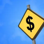 A yellow road sign with a dollar sign in black. Sits to the right of the image on a pole, with a blue sky background.