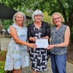 Three ladies stand together in a garden, holding a cheque.