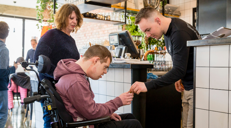 A person stands behind a person in a wheelchair, watching on as they receive change from a waiter