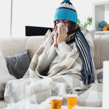 A person sits on a couch wearing a beanie and wrapt in a blanket. They are blowing their nose. Medicine sits on the coffee table in front.