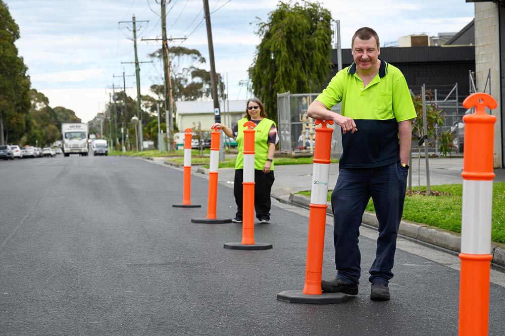 Two people standing on road in high vis, leaning against traffic bollards on the road.