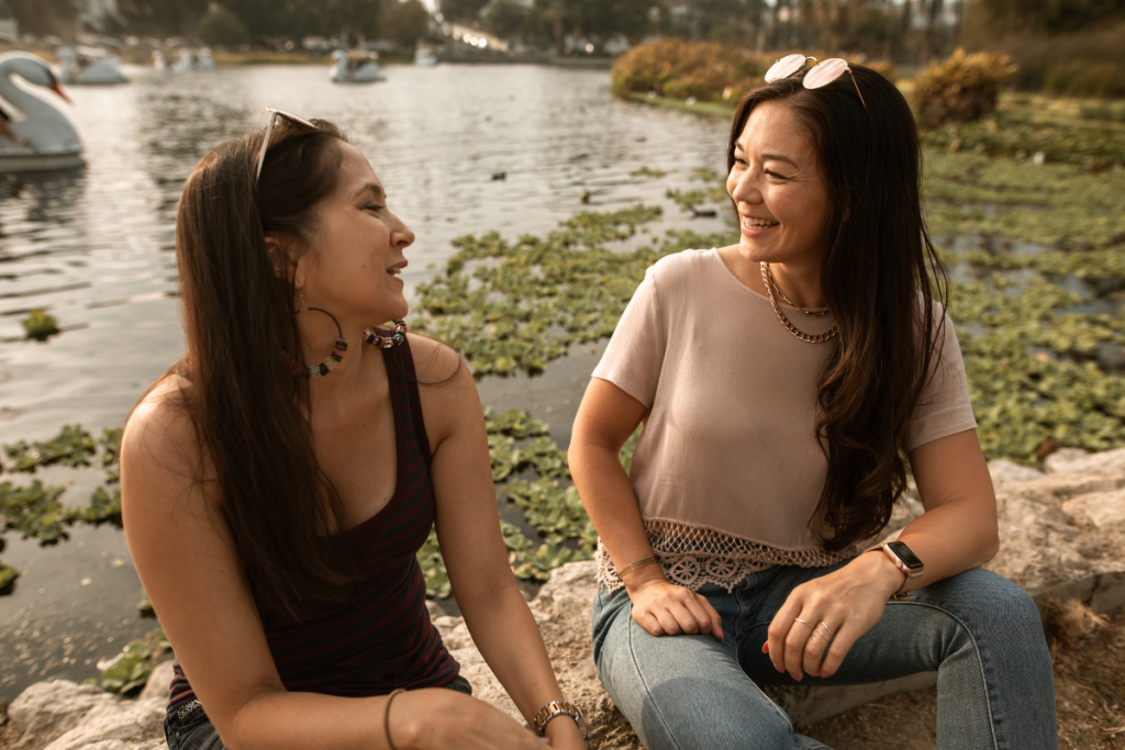 Two women sit next to each other outdoors, talking and smiling.