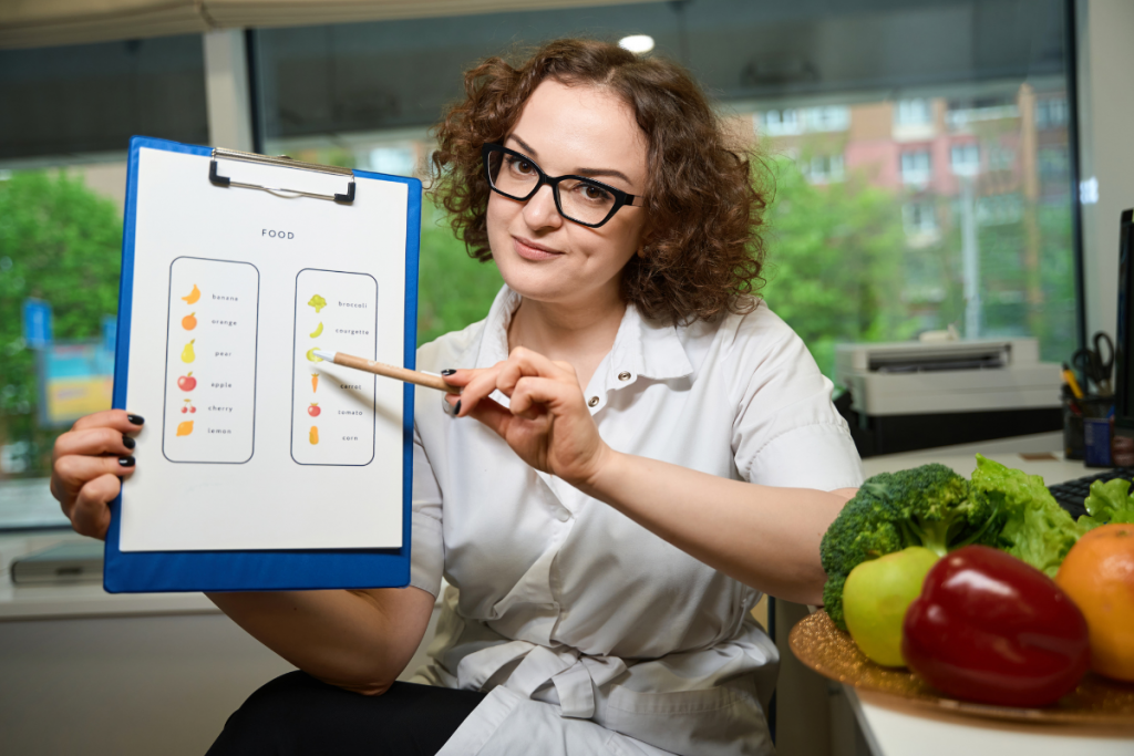 Woman sitting near some fresh vegetables holds up a clipboard with a visual list of vegetables.